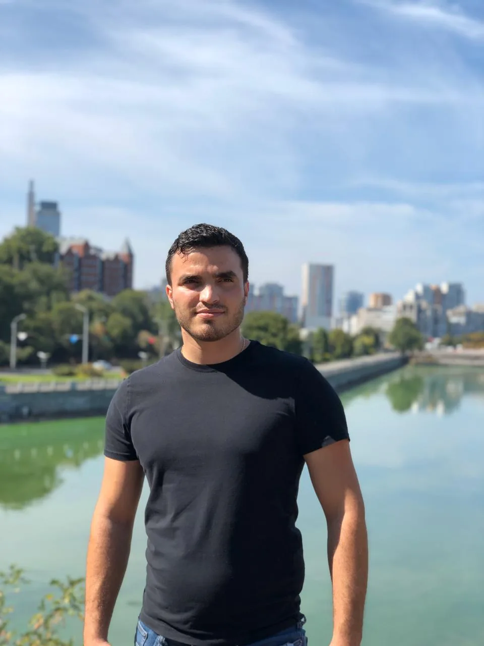 Edward Musaelyan, in a black shirt, standing outdoors with a river and a city skyline behind him